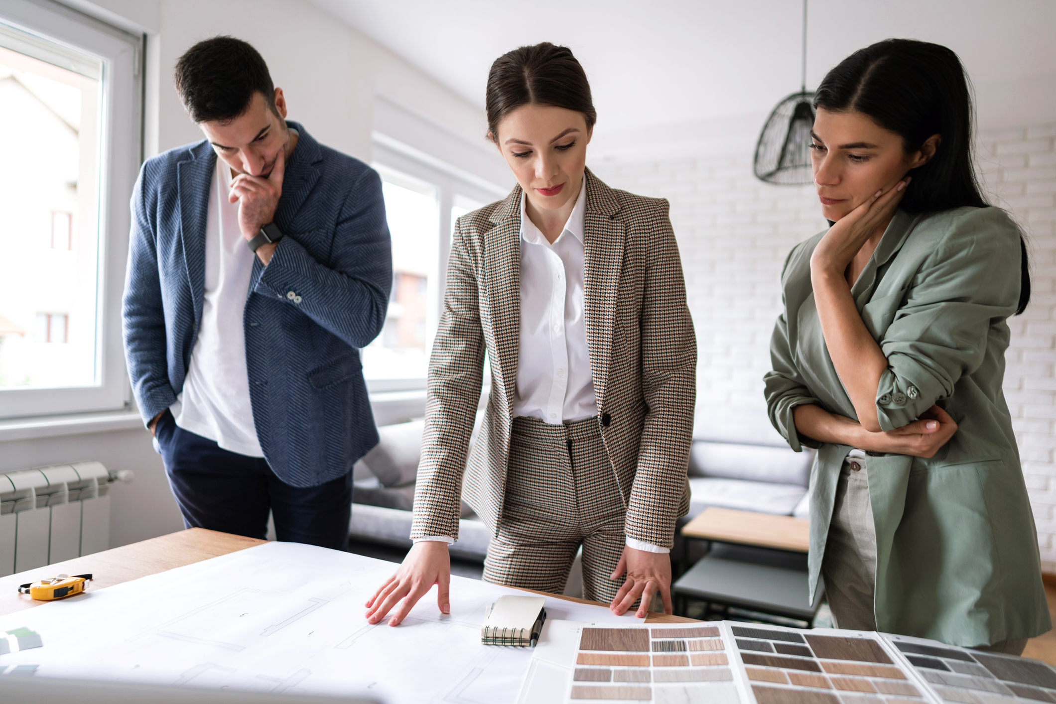 Stock Image of a Real Estate Agent Standing at a Table Looking Down At House Plans and Color Selections While a Man and Woman Stand on Either Side of Her with Their Arms Crossed As If in Disagreement With the Agent