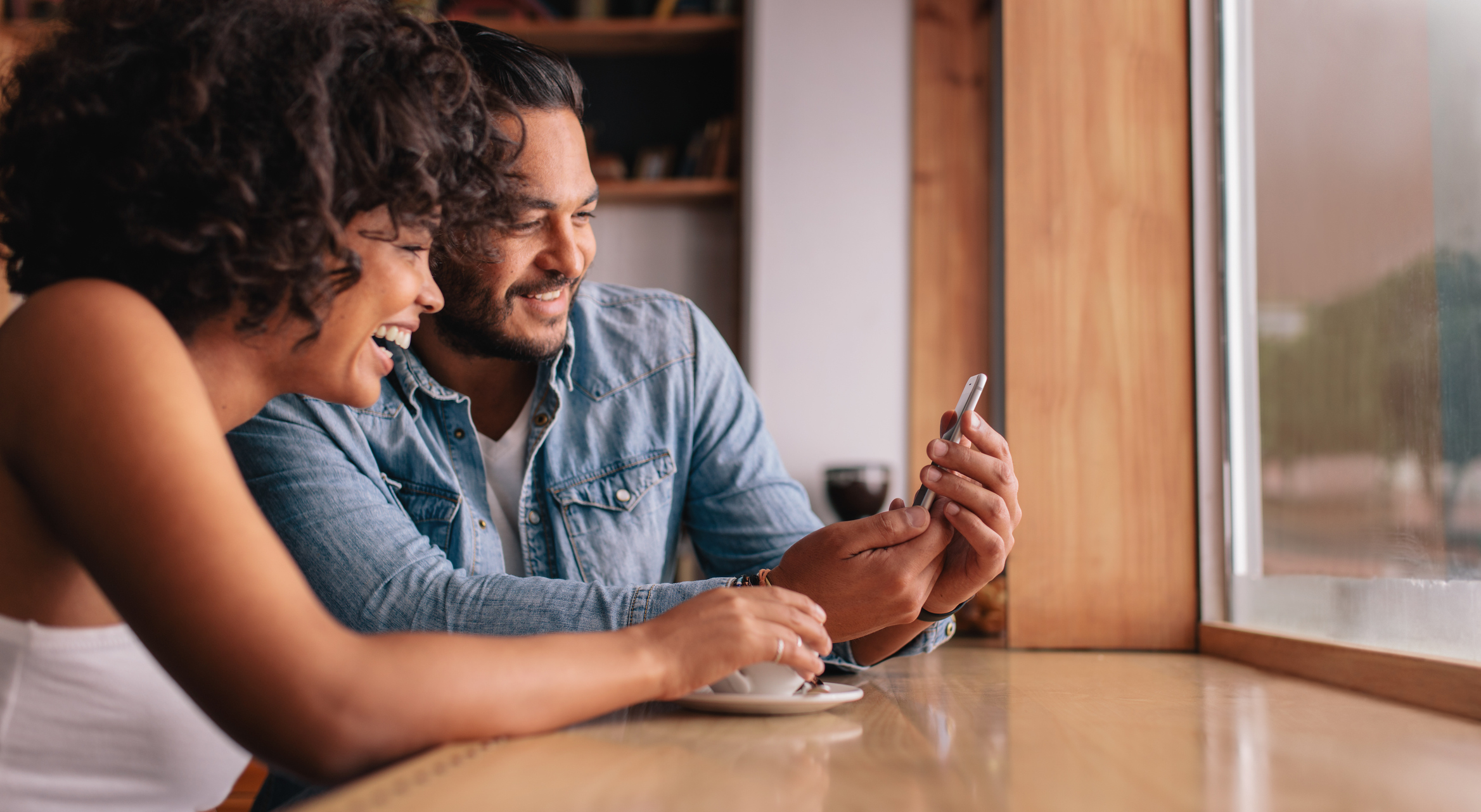 Stock Image of a Woman Holding A Cup Of Coffee and Man Holding A Cell Phone Sitting Together at A Table and Laughing At What Is on the Cell Phone