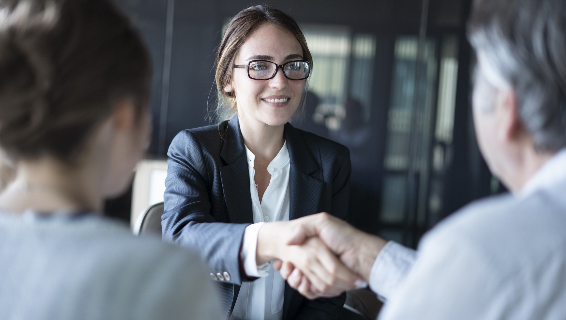 Stock Image of a Woman in A Business Suit and Glasses Smiling While Extending Her Arm and Shaking Hands with A Person Out Of View