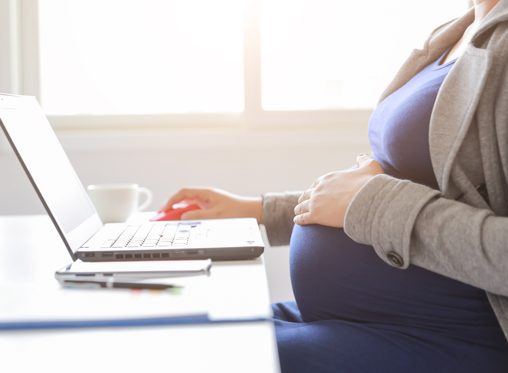 Stock Image of Pregnant Business Woman Sitting at Desk with One Hand on Her Pregnant Belly and the Other Hand on Laptop Mouse. We Cannot See Her Face But It Appears She Is Working on the Laptop