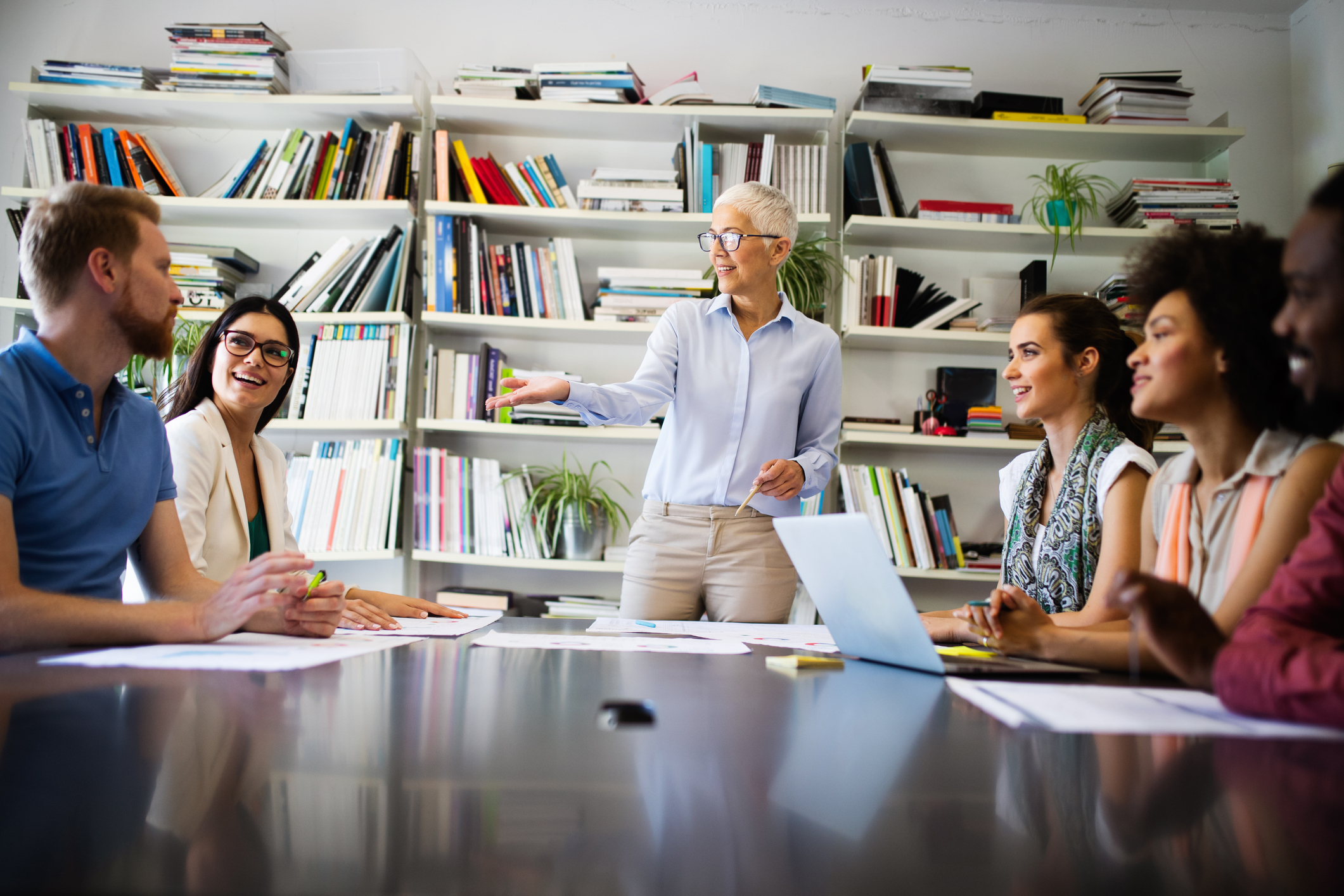 Stock Image of Team Meeting Older Woman Is Standing at the Head of the Table Explaining Something To Five Individuals Around the Table