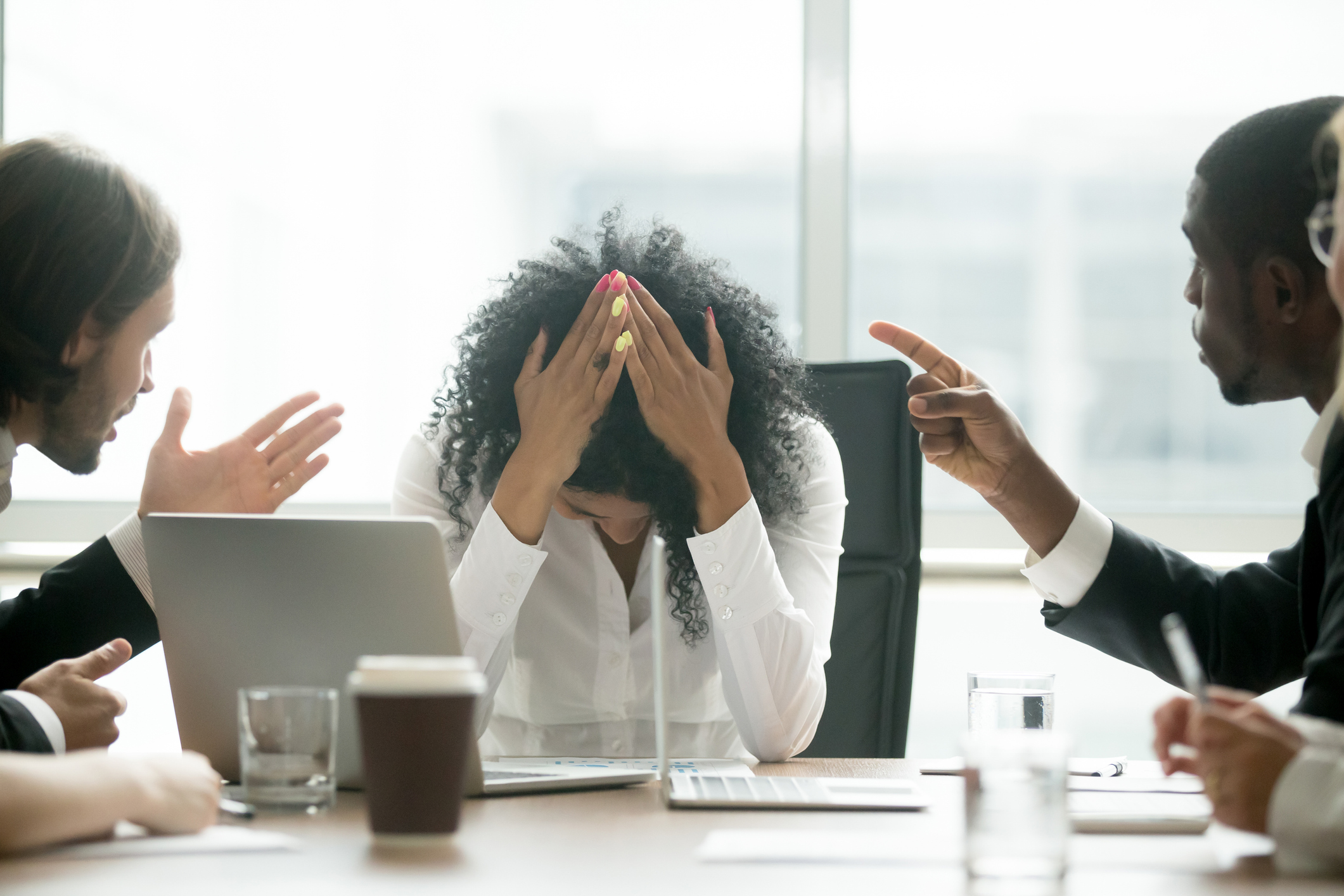 Stock Photo of a Woman Sitting at a Conference Table with Her Head in Her Hands and Two Men on Either Side of Her Pointing At Her and Appearing To Yell