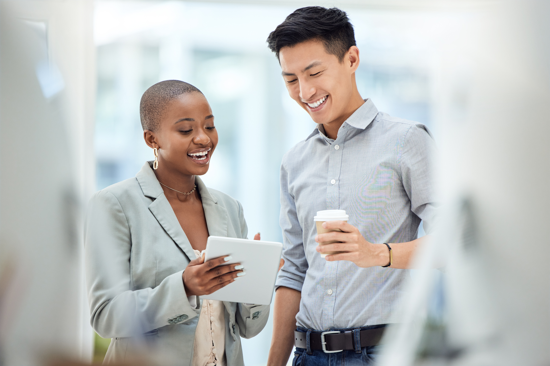 Stock image of a man and woman standing in an office hallway. The man is holding a coffee and smiling, and the woman is smiling and looking at a report in her hands. 