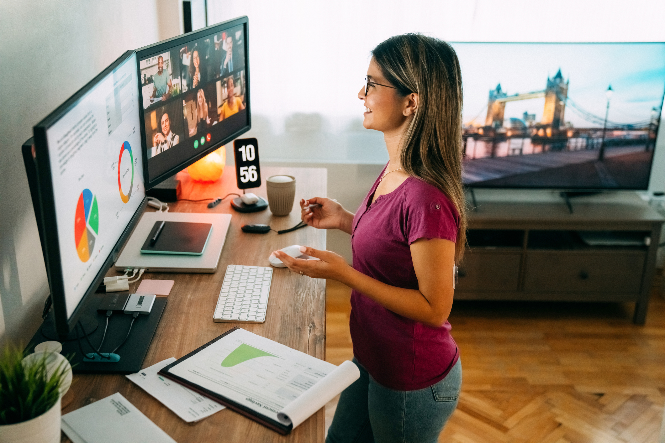 Stock image of woman on zoom meeting standing in front of two computer monitors and standing at a standing desk in a living room.