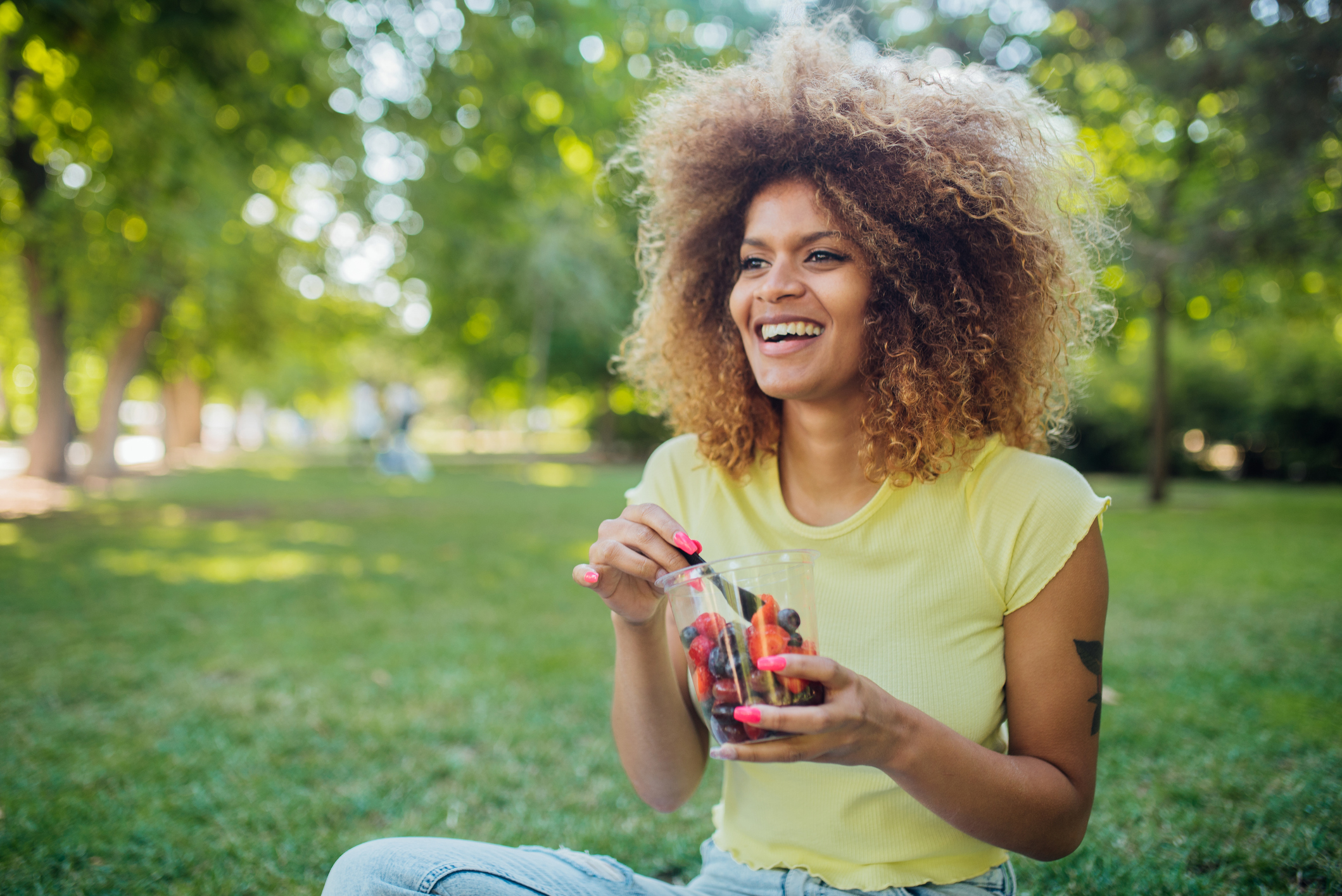 Stock image of a woman sitting on the grass in a park and eating a bowl of fruit while smiling and looking in the distance. 