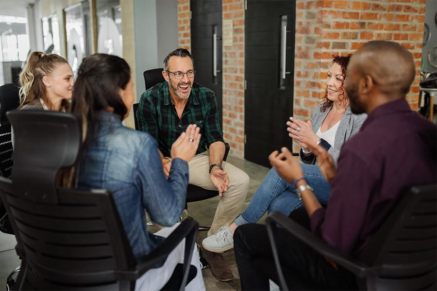 five business colleagues sitting in a circle in an office and smiling and talking with each other