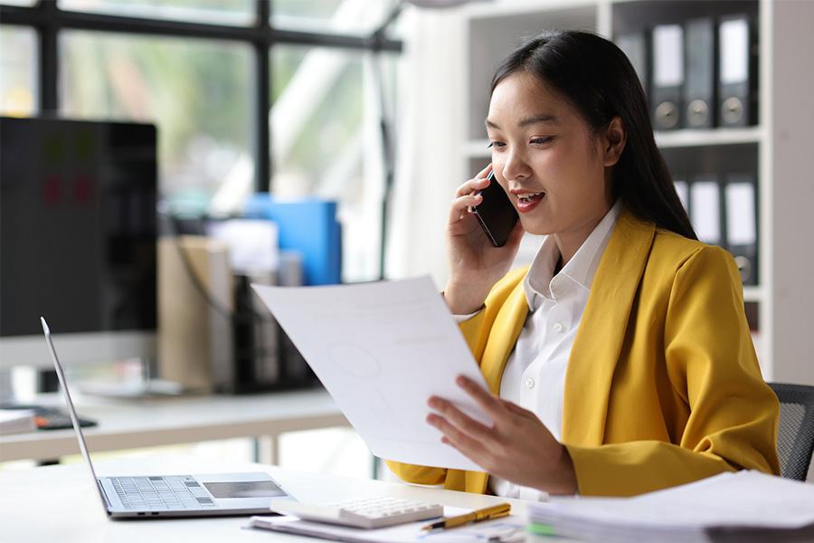 business woman sitting in an office in front of her laptop and holding a piece of paper while on her cell phone