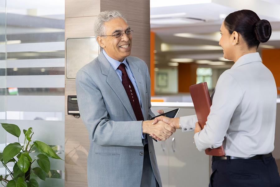 older business man standing in an office hallway smiling and shaking hands with a younger business woman
