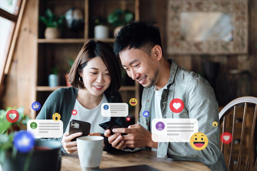 man and woman sitting at a kitchen table drinking coffee and looking at their cell phones. both are smiling and the woman is glancing at the man's phone screen