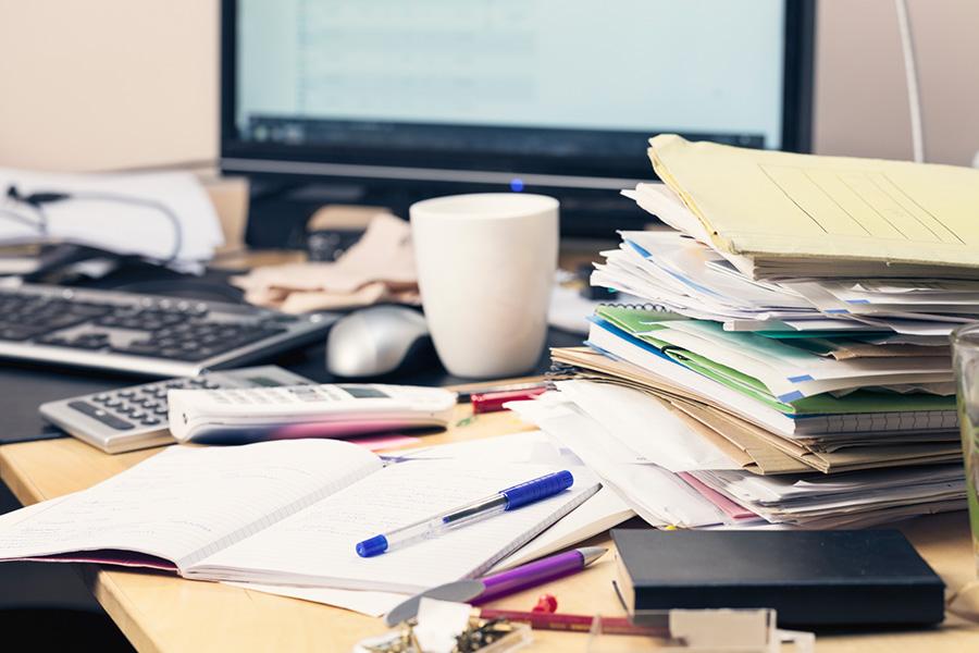 close up image of a disorganized work desk with several piles of paper and folders, a coffee cup, calendar and open notebook. 