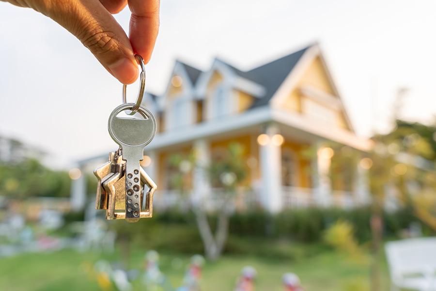 stock image of big, white house blurred in the background and a persons hand extending in front of the camera holding a set of keys