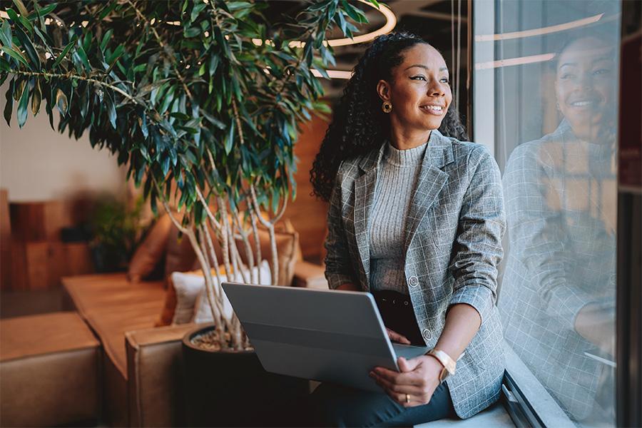 business woman sitting by window in a chic office, looking out the window with a laptop on her lap and smiling 