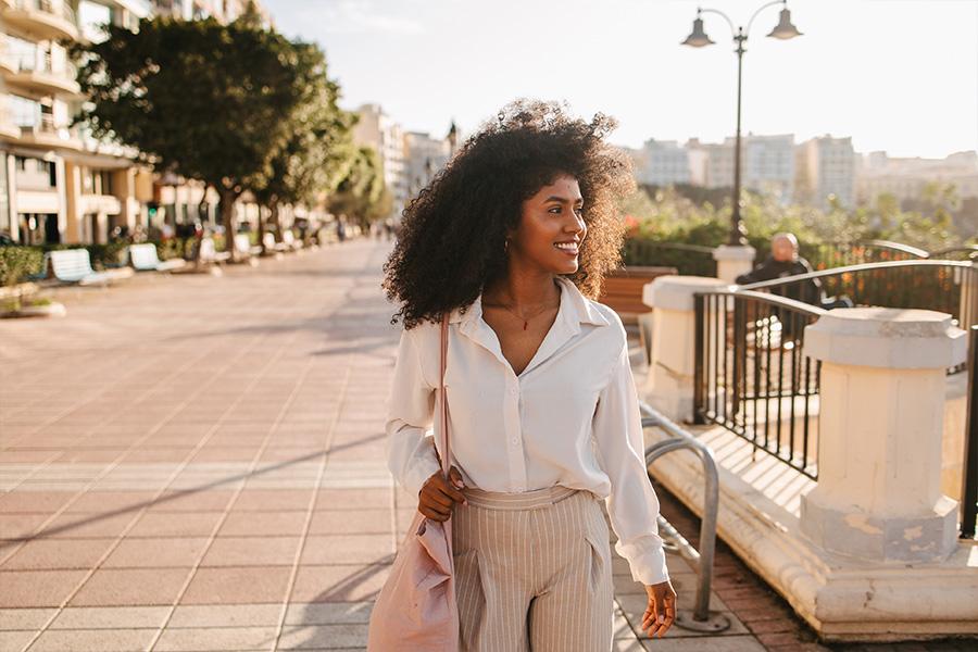 stock image of a woman walking down a pedestrian path near a row of buildings, her hair is blowing and she is smiling looking away from the camera