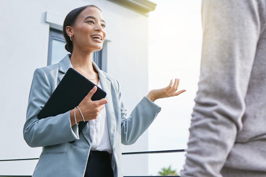 stock image of woman standing in front of a house and speaking to someone holding a tablet in one arm
