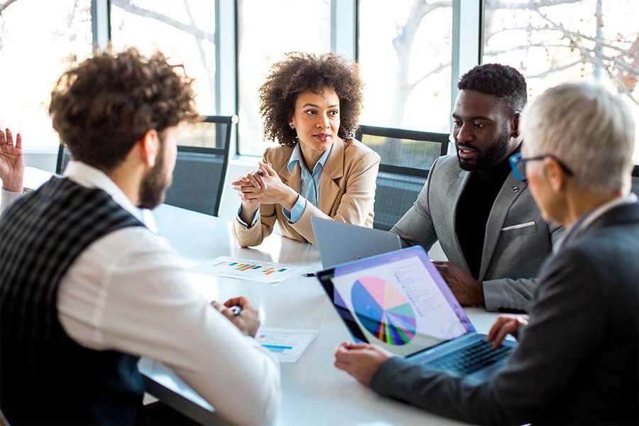 stock image of two men and two women at a business meeting, sitting at a table with one woman looking at a computer screen
