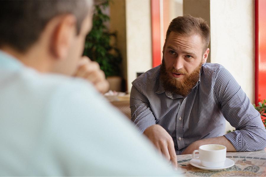 stock image of two men having a conversation at a table with coffee. one man is in deep thought speaking to the other