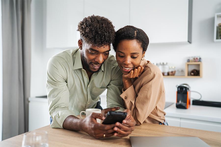 stock image of a young couple leaning on their kitchen counter smiling and looking at a cell phone screen that is in the mans hands