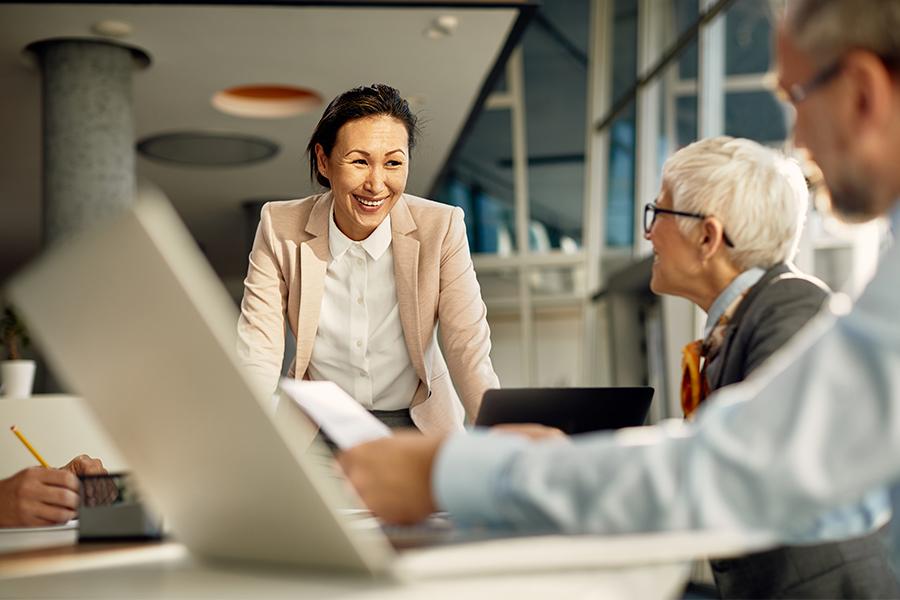 stock image of two people sitting at a work table and a woman is standing beside them leaning on table smiling